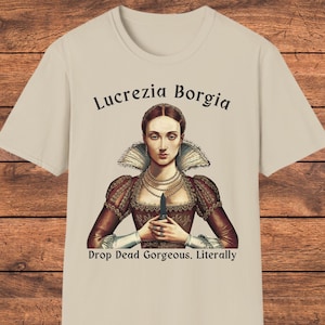 May include: A beige t-shirt featuring a graphic of Lucrezia Borgia wearing a dark red dress with a high collar and pearl necklaces, holding a small dagger. The text reads Lucrezia Borgia above and Drop Dead Gorgeous. Literally below the figure.