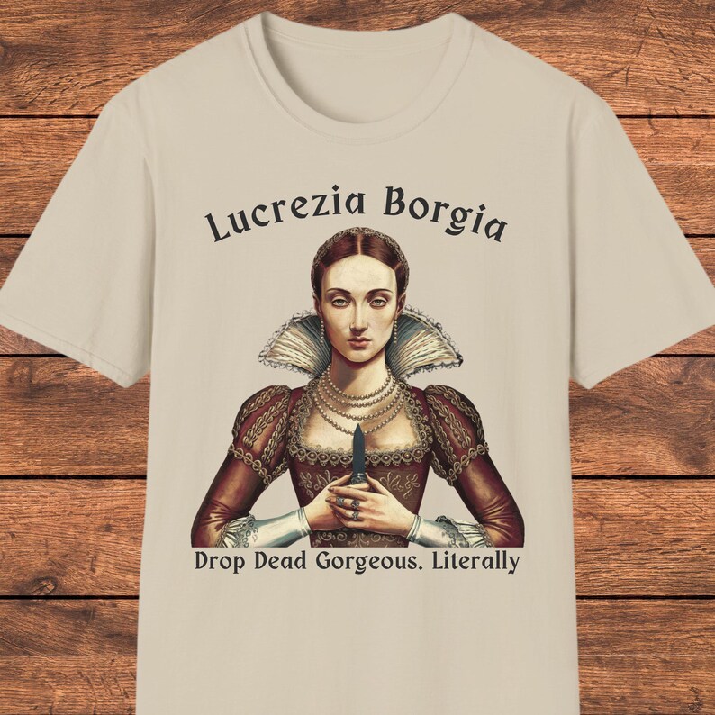 May include: A beige t-shirt featuring a graphic of Lucrezia Borgia wearing a dark red dress with a high collar and pearl necklaces, holding a small dagger. The text reads Lucrezia Borgia above and Drop Dead Gorgeous. Literally below the figure.