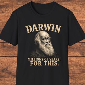 Könnte beinhalten: Ein schwarzes Kurzarm-T-Shirt mit einer beigen Grafik von Charles Darwin mit langem Bart. Der Text DARWIN ist in einer verwaschenen beigen Schrift über dem Porträt gedruckt, und der Text MILLIONS OF YEARS. FOR THIS. steht darunter.