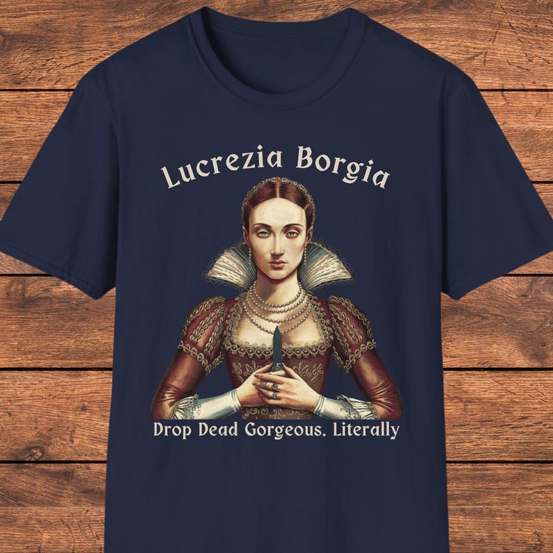 May include: A navy blue t-shirt featuring a graphic illustration of Lucrezia Borgia in a red dress with a high collar and pearl necklaces, holding a small dagger. The text reads Lucrezia Borgia above and Drop Dead Gorgeous. Literally below.