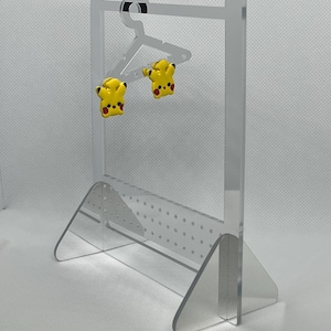 May include: Clear acrylic earring display stand with a hanger-shaped top and a perforated base. Two yellow and red Pikachu earrings are displayed on the hanger. The stand has a modern, minimalist design.