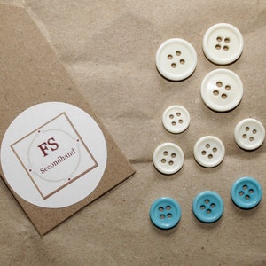 Könnte beinhalten: Ein braunes Papieretikett mit dem Text "FS Secondhand" auf einem weißen runden Aufkleber. Es sind neun Knöpfe auf dem Bild zu sehen, sechs weiße Knöpfe mit jeweils vier Löchern und drei blaue Knöpfe mit jeweils sechs Löchern.