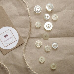 Könnte beinhalten: Eine Sammlung von 12 cremefarbenen Plastikknöpfen mit jeweils zwei Löchern. Die Knöpfe sind auf einem braunen Papierhintergrund mit einem braunen Papieretikett mit der Aufschrift "FS Secondhand" angeordnet.