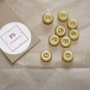 Könnte beinhalten: Zehn runde, cremefarbene Knöpfe mit jeweils zwei Löchern. Die Knöpfe sind auf einer braunen Papieroberfläche angeordnet.