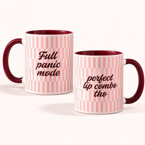 May include: Two ceramic mugs with a pink and white vertical striped pattern. The left mug features the dark red text Full panic mode, while the right mug displays the dark red text perfect lip combo tho. Both mugs have a dark red interior and handle.