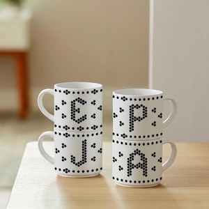 May include: Four white ceramic mugs with handles, arranged in two stacks. Each mug features a black letter E, J, P, or A formed by a honeycomb pattern. The design includes horizontal rows of black dots and small hexagonal shapes encircling the mugs.