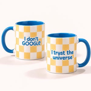 May include: Two ceramic mugs with a yellow and white checkered pattern. One mug features the blue text "I don't GOOGLE" and the other features the blue text "I trust the universe". Both mugs have a solid blue interior and a blue handle.