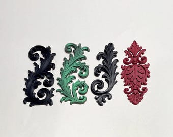 May include: Four decorative scrollwork appliques featuring intricate leaf and vine patterns. The set includes two black pieces, one green piece with dark shading, and one deep red piece, all displaying raised relief details and curved edges.