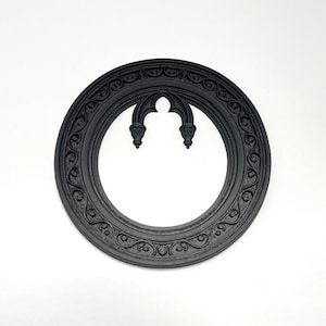May include: A black, circular decorative frame with an ornate, gothic-style design. The frame features a repeating scroll pattern around the outer edge and a stylized arch at the top. The frame is made of a solid material and is set against a white background.