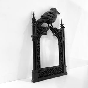 Puede incluir: Marco decorativo negro de estilo g&oacute;tico con un cuervo posado. El marco tiene una parte superior arqueada y detalles ornamentados. El cuervo a&ntilde;ade un toque g&oacute;tico. El marco est&aacute; hecho de un material oscuro.