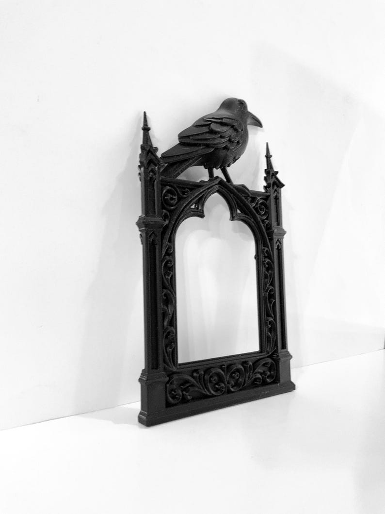 Puede incluir: Marco decorativo negro de estilo g&oacute;tico con un cuervo posado. El marco tiene una parte superior arqueada y detalles ornamentados. El cuervo a&ntilde;ade un toque g&oacute;tico. El marco est&aacute; hecho de un material oscuro.