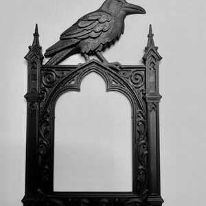 Puede incluir: Marco g&oacute;tico negro con un cuervo decorativo en la parte superior. El marco tiene una abertura arqueada y detalles ornamentales. El cuervo est&aacute; detallado con plumas texturizadas y un pico prominente. Dise&ntilde;o oscuro y g&oacute;tico.