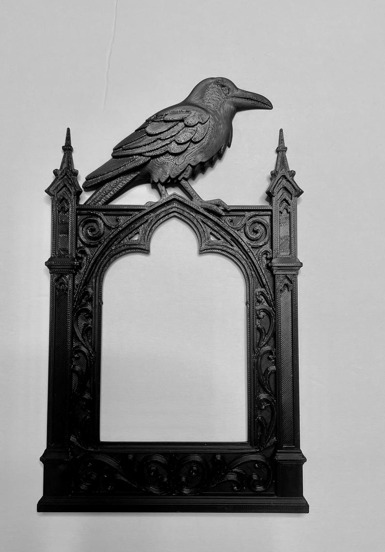 Puede incluir: Marco g&oacute;tico negro con un cuervo decorativo en la parte superior. El marco tiene una abertura arqueada y detalles ornamentales. El cuervo est&aacute; detallado con plumas texturizadas y un pico prominente. Dise&ntilde;o oscuro y g&oacute;tico.