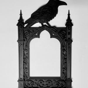 Puede incluir: Marco de fotos g&oacute;tico negro con un cuervo posado encima. El marco tiene una abertura arqueada y detalles ornamentados. El cuervo est&aacute; esculpido en una pose realista. El marco est&aacute; hecho de un material oscuro, posiblemente madera o resina.
