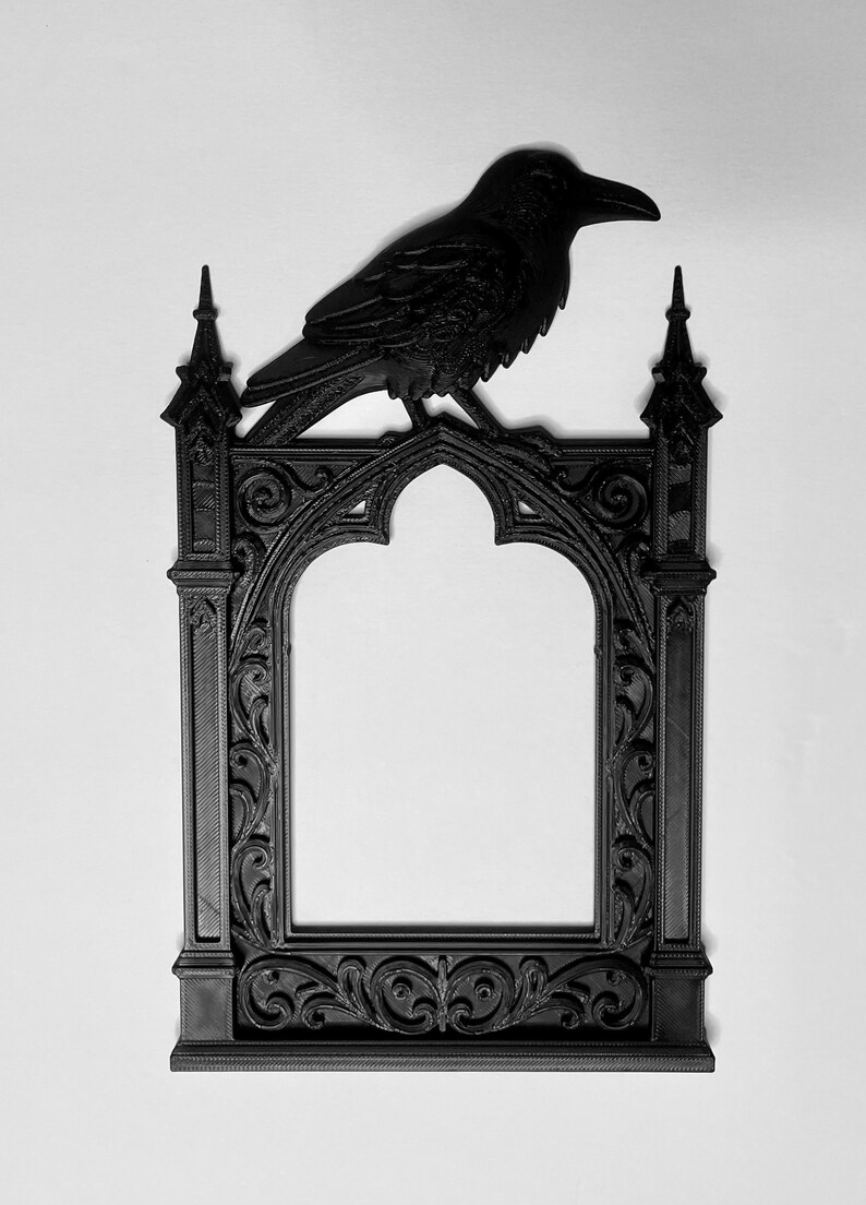 Puede incluir: Marco de fotos g&oacute;tico negro con un cuervo posado encima. El marco tiene una abertura arqueada y detalles ornamentados. El cuervo est&aacute; esculpido en una pose realista. El marco est&aacute; hecho de un material oscuro, posiblemente madera o resina.