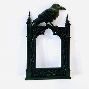 Puede incluir: Un marco decorativo verde oscuro con un dise&ntilde;o de arco g&oacute;tico, coronado por una detallada escultura de cuervo. El marco presenta adornos ornamentados y pin&aacute;culos puntiagudos, creando una est&eacute;tica vintage.