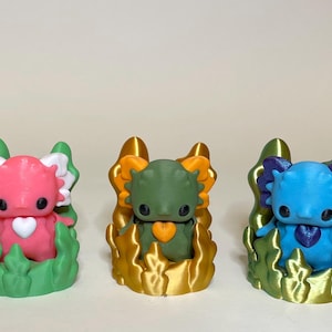 May include: Three small, colorful figurines of axolotls, each with a different colored body and a heart-shaped design on their chest. The figurines are sitting on bases made of green, gold, and blue plastic, resembling aquatic plants.