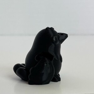 May include: A black 3D printed figurine of a bird with its wings folded. The bird is sitting on a white surface.