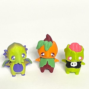 May include: Three colorful figurines on a white background. The figurines include a green creature with purple accents, an orange pumpkin-like figure, and a green zombie-like figure with a skull on its chest. The figures have large eyes.