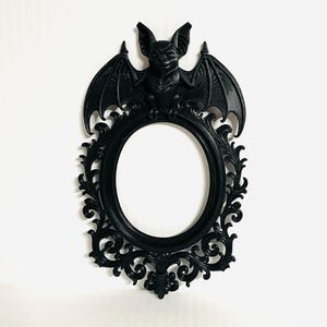 May include: Black oval-shaped picture frame with a bat on top. The frame has ornate, swirling details and a smooth, glossy finish. The bat has its wings spread and is centered above the frame. The frame is designed to be hung on a wall.