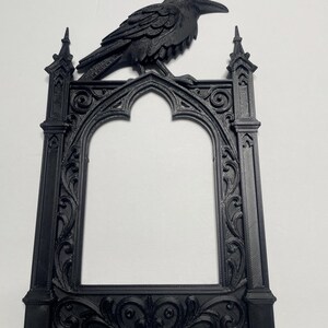 Puede incluir: Un marco de fotos g&oacute;tico negro con un cuervo en la parte superior. El marco presenta adornos de volutas y un dise&ntilde;o de arco apuntado. El cuervo est&aacute; intrincadamente esculpido, agregando un encanto g&oacute;tico. Un art&iacute;culo decorativo.