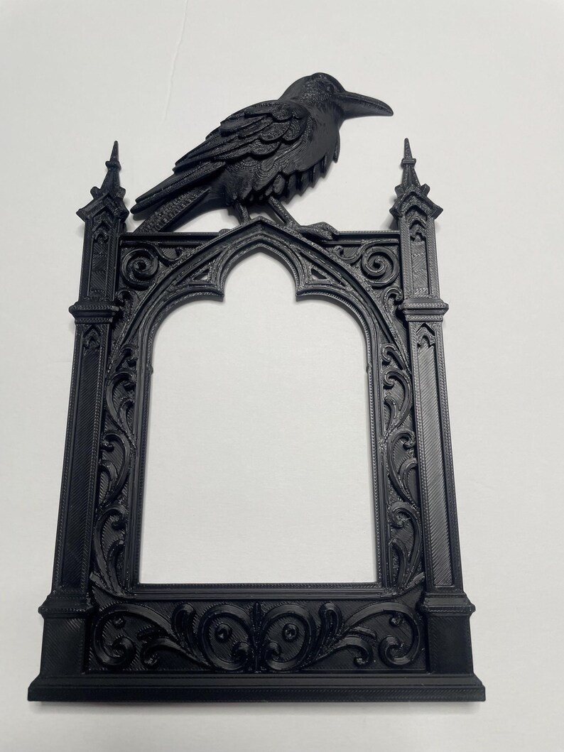 Puede incluir: Un marco de fotos g&oacute;tico negro con un cuervo en la parte superior. El marco presenta adornos de volutas y un dise&ntilde;o de arco apuntado. El cuervo est&aacute; intrincadamente esculpido, agregando un encanto g&oacute;tico. Un art&iacute;culo decorativo.