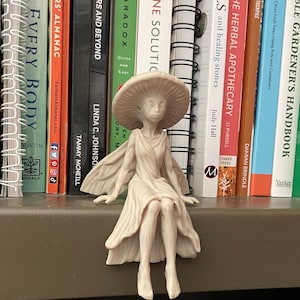 May include: A light beige figurine of a humanoid creature with a large mushroom cap for a head, delicate wings, and a flowing dress. The figure is depicted in a seated position with legs dangling, featuring intricate sculpted details on the cap gills.