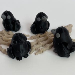 May include: Three small black crow figurines with gray eyes, sitting on a white skeletal hand prop.