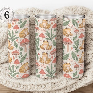 May include: Three stainless steel tumblers with a cream background and a repeating pattern of green leaves, red mushrooms, and yellow frogs. The tumblers are on a white knitted blanket.