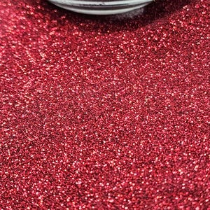 May include: Close-up of a vibrant red glitter, with a glass container partially visible at the top. The glitter is densely packed, creating a sparkling, textured surface. Ideal for crafts, decorations, or adding shimmer to various projects.