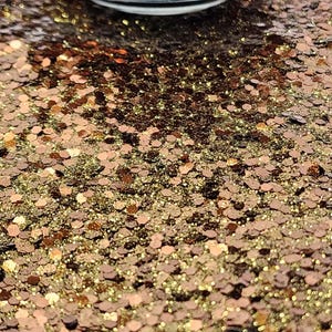 May include: Close-up of a surface covered in copper and gold glitter. The glitter is in various shapes and sizes, reflecting light. A clear glass container is partially visible at the top of the image.