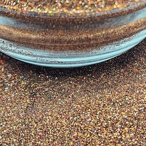 May include: Close-up of a glass container filled with shimmering brown glitter. The glitter has a metallic sheen with hints of gold and copper. The container has a clear, rounded rim, and the glitter is densely packed.