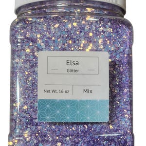 May include: A clear plastic jar filled with Elsa glitter mix. The glitter is a mix of purple, blue, and iridescent particles. The jar has a white lid and a label that reads "Elsa Glitter" and "Net Wt. 16 oz."