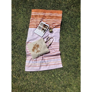 Puede incluir: Una toalla de playa con rayas horizontales en tonos naranja, blanco y rosa, con un bolso tote a juego con un diseño celestial. Una revista y un vaso están sobre la toalla, colocados sobre un césped verde.