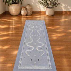 May include: A rectangular yoga mat in a muted blue shade featuring a white symmetrical print of two winding snakes, celestial stars, and crescent moon motifs. The design is enclosed within a decorative border of geometric patterns and small dots.