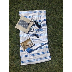 Puede incluir: Una toalla de playa a rayas blancas y azules con una bolsa de mano azul, una revista, una cámara azul, gafas de sol y una botella de champán en la parte superior. La toalla tiene la palabra "Marbella" escrita en ella.
