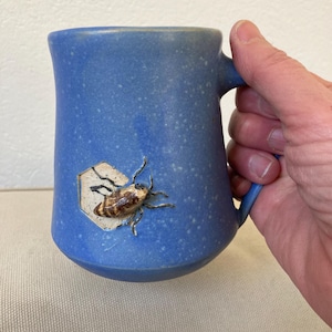 Op de afbeelding: Een blauwe keramische mok met een getextureerd oppervlak. De mok heeft een handvat en een reliëfdesign van een bij aan de voorkant.