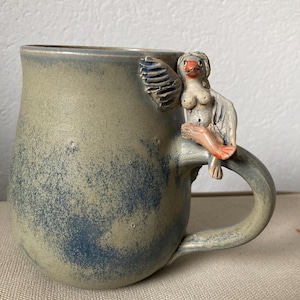 Könnte beinhalten: Eine handgefertigte Keramik-Tasse mit einem einzigartigen Design. Die Tasse ist hauptsächlich in gedämpftem Grün mit blauen Akzenten gehalten. Eine geformte Figur einer Vogel-Frau ziert den Griff. Die Vogelfrau hat weiße Flügel, einen roten Schnabel und orangefarbene Füße. Ein einzigartiges Kunstwerk.
