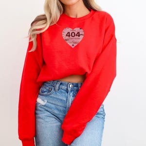 Op de afbeelding: Een felrood sweatshirt met een hartvormige afbeelding. De afbeelding toont de tekst "ERROR 404 LOVE NOT FOUND" in een distressed stijl. Het sweatshirt is effen van kleur en heeft lange mouwen.
