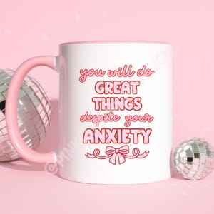 May include: White ceramic mug with a pink handle and interior. The mug features the text "you will do GREAT THINGS despite your ANXIETY" in pink and red lettering. A pink bow graphic is at the bottom. The background is pink with disco balls.