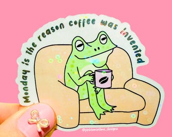 May include: A green frog sitting on a sofa with a cup of coffee. The text on the sticker reads "Monday is the reason coffee was invented."