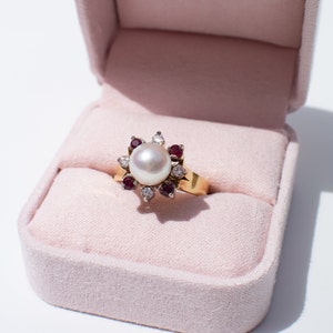 May include: A gold ring featuring a large white pearl surrounded by smaller diamonds and red rubies. The ring is displayed in a pink velvet ring box.