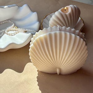Può includere: Una collezione di candele a forma di conchiglia e un piatto decorativo. Le candele color avorio, di cui una accesa, hanno una consistenza a coste. Un piatto bianco con effetto marmorizzato blu contiene una collana di perle. Tema costiero.