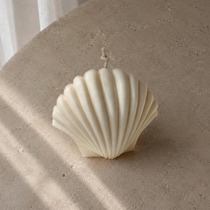 Può includere: Una candela a forma di conchiglia color crema con una superficie strutturata. La candela ha uno stoppino in cima ed è posta su uno sfondo neutro. Il design della candela imita la forma naturale di una conchiglia.