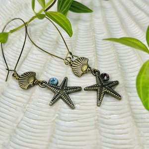 May include: A pair of bronze-toned earrings featuring a seashell design at the top and a starfish pendant. One earring has a blue gem, the other a purple gem. The earrings are displayed on a white, textured surface with green foliage.