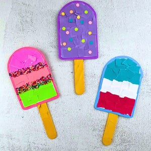 May include: Three colorful popsicle crafts. One is purple with colorful dots, another is pink, coral, and green with sprinkles, and the last is blue, white, and red. Each has a yellow popsicle stick.