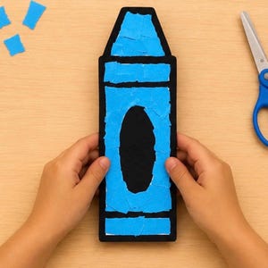 May include: A black crayon-shaped cutout decorated with torn pieces of bright blue paper. The central portion of the crayon features a large, solid black oval shape. The cutout is positioned vertically, with small blue paper scraps nearby.