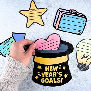 May include: A collection of colorful New Year's goals cards. The cards are shaped like a star, suitcase, heart, lightbulb, and a top hat with the text "NEW YEAR'S GOALS!". The heart-shaped card reads "I'll spread kindness by..."