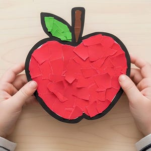 Puede incluir: Una figura de manzana hecha de papel con un contorno negro, un tallo marrón y una hoja verde. El cuerpo principal de la manzana está relleno con un mosaico de trozos de papel rojo rasgado, creando una superficie texturizada dentro del borde.