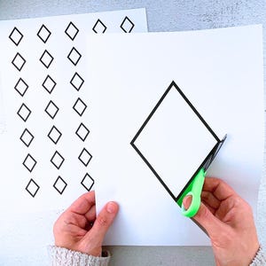 Könnte beinhalten: Zwei weiße Papierbögen mit schwarzen Rautenformen bedruckt. Ein Bogen hat mehrere kleine Rauten, der andere eine große Raute, die mit einer grün-griffigen Schere ausgeschnitten wird. Die Schere schneidet die große Rautenform aus.